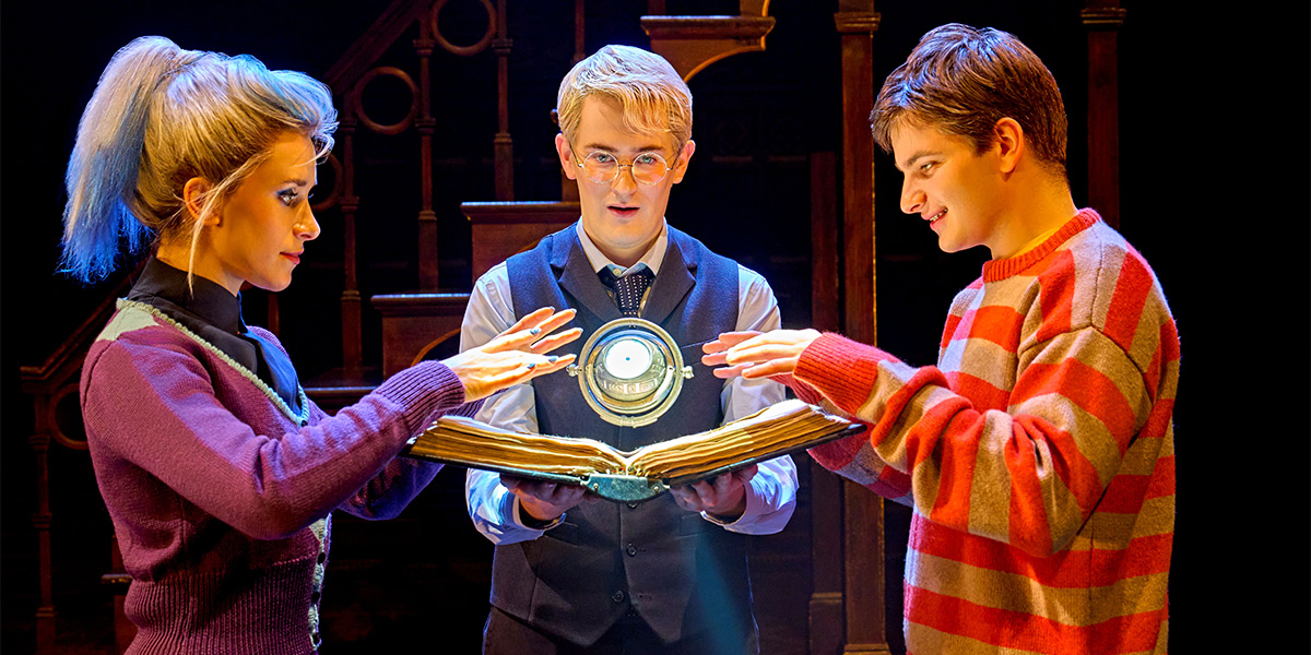 Five young adults in wizarding attire stand together, smiling, with a backdrop of a magical setting.