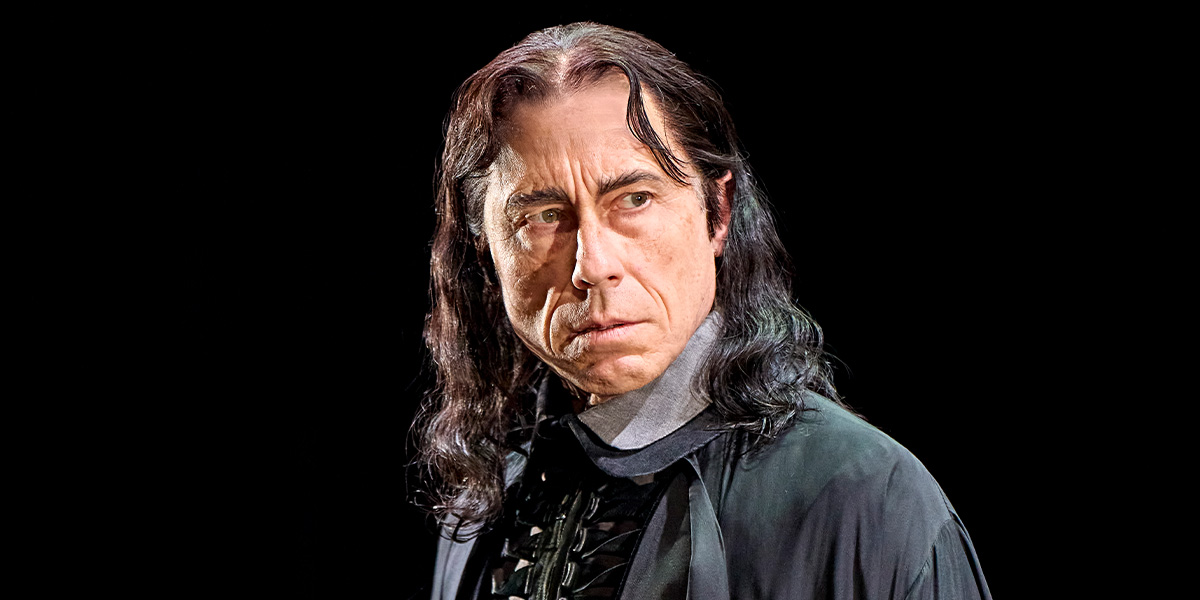 David Annen as Severus Snape, wearing a black robe, stands with a serious expression against a dark background.
