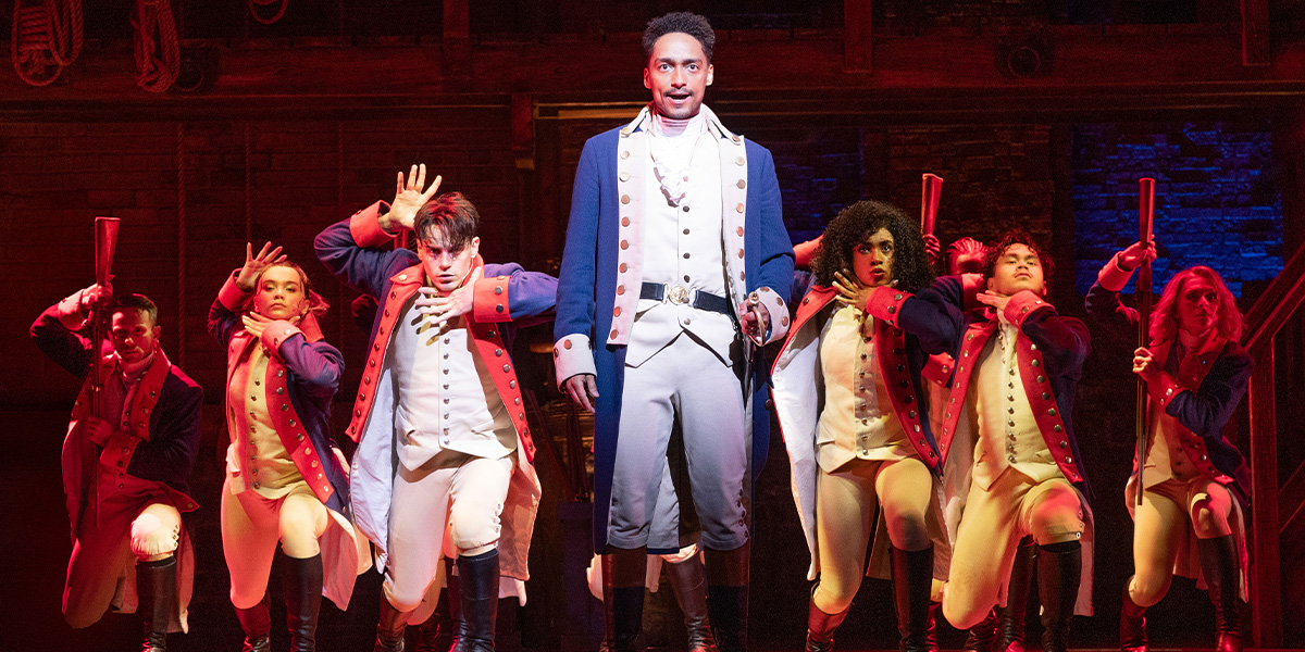 Alex Sawyer as Alexander Hamilton, wearing a period costume, stands confidently with arms crossed against a dark background.