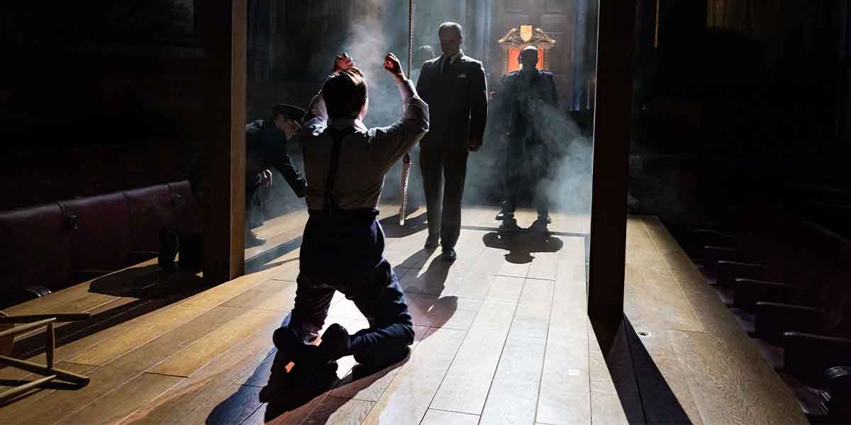 A kneeling actor in suspenders gestures dramatically, while two figures stand in the background, shrouded in smoke.
