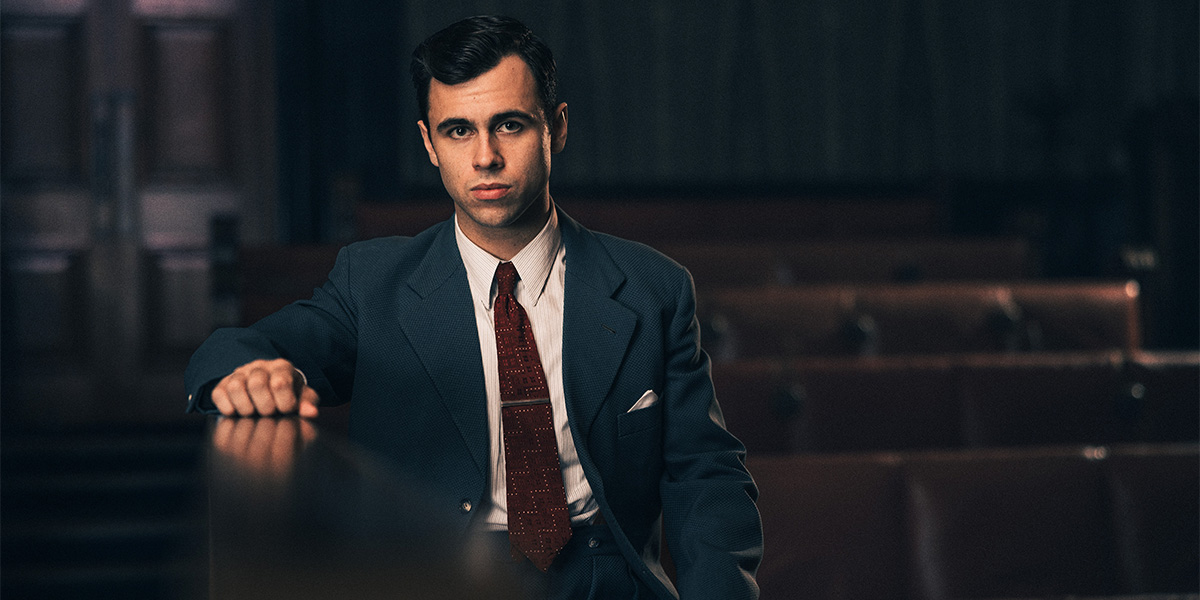 A man in a blue suit and red tie sits pensively at a courtroom railing, with empty seats behind.