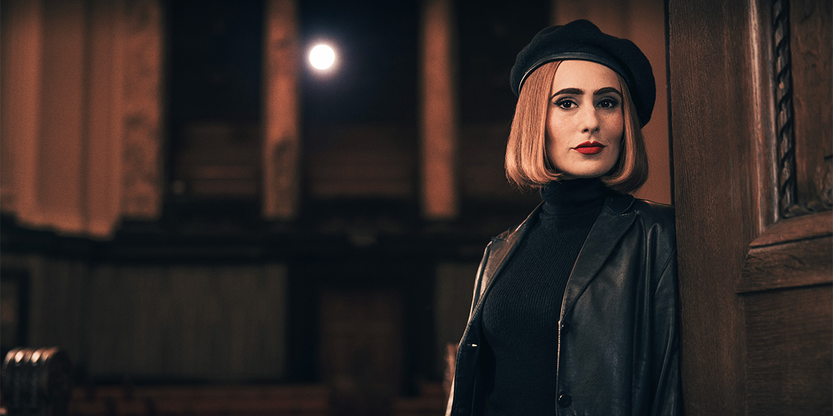 A woman with a bob haircut and red lipstick leans against a wooden wall, wearing a black beret and leather jacket.