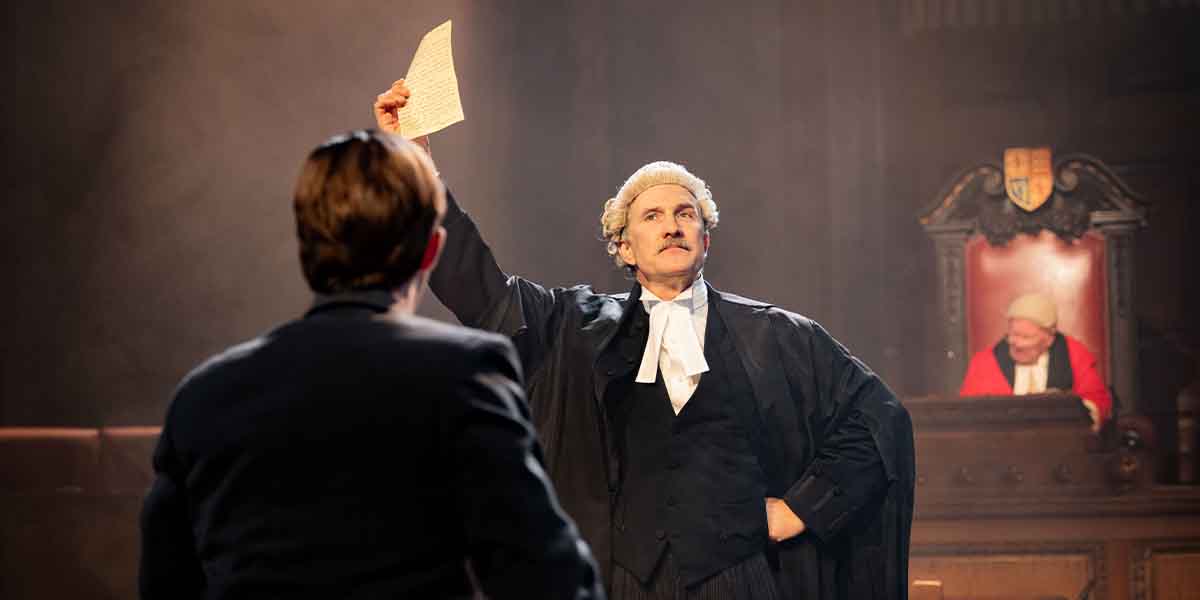 Matthew McGiven as Leonard Vole holds a document aloft, while Jo Stone-Fewings observes from the background.