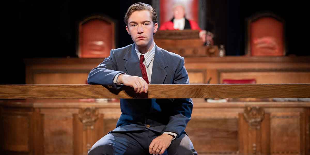 Matthew McGivern as Leonard Vole sits at a wooden witness stand in a courtroom setting.