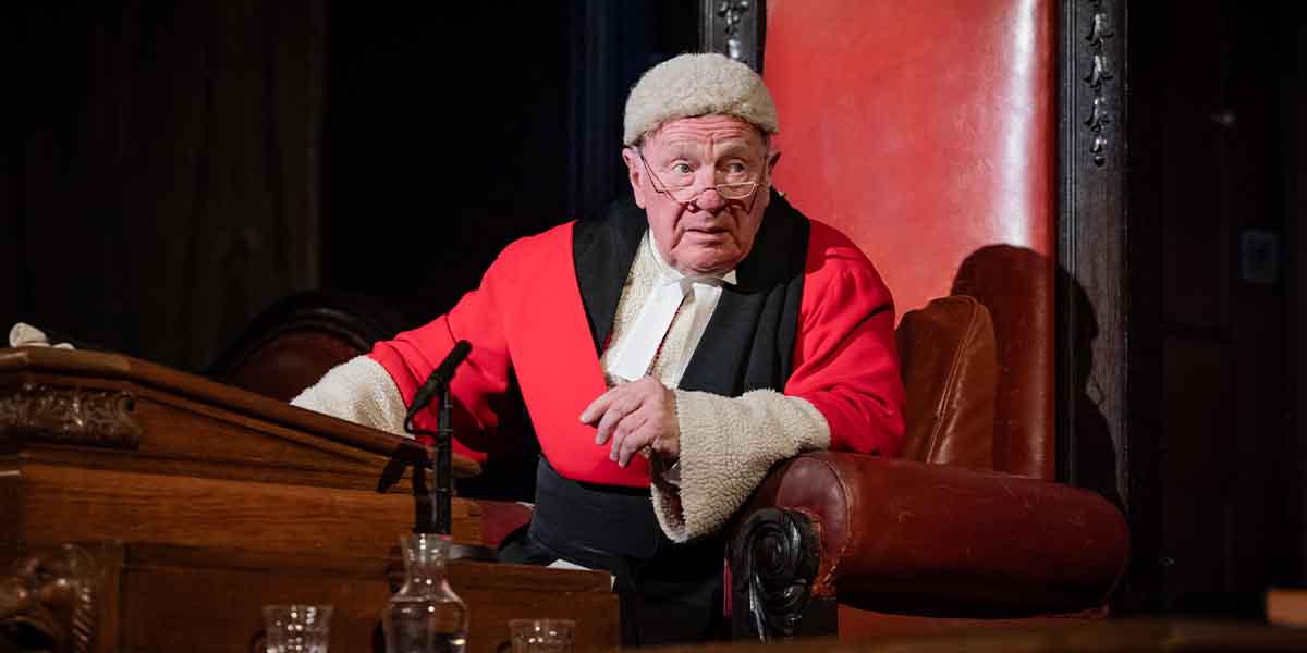 Mr Justice Wainwright, in a red robe and wig, sits at a wooden desk, looking intently.