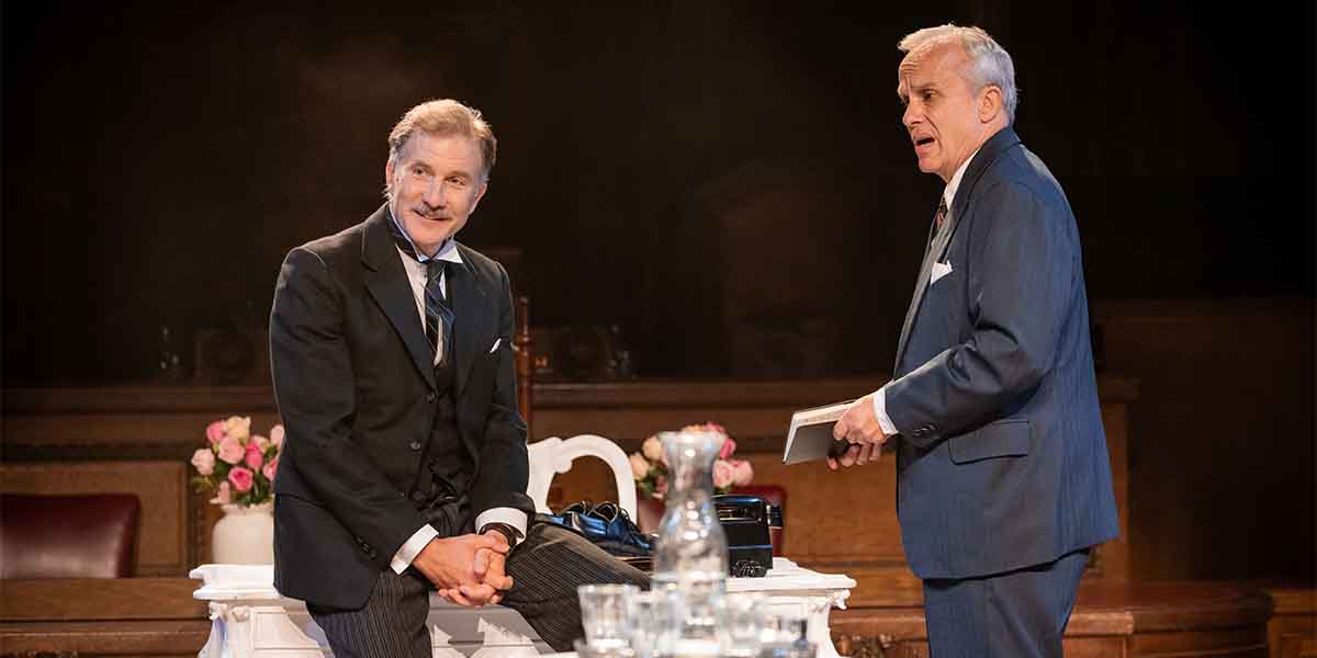 Jo Stone-Fewings as Sir Wilfrid Robarts sits on a table, while Crispin Redman as Mr Mayhew stands nearby.
