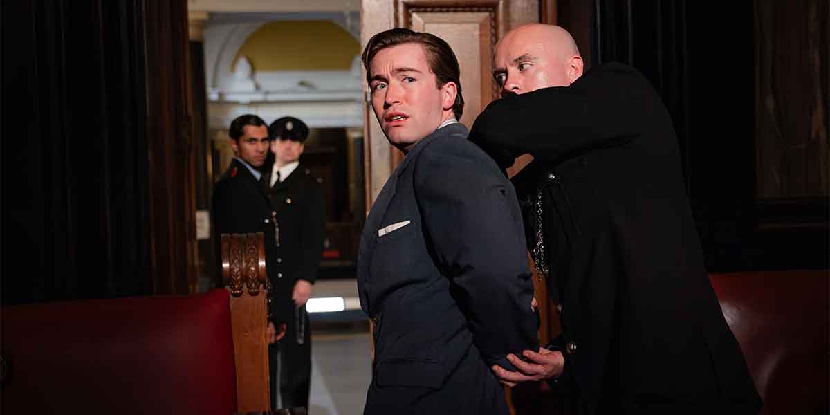 Matthew McGiven as Leonard Vole is being restrained by Sean Paul Jenkinson as Sergeant Randall in a courtroom.