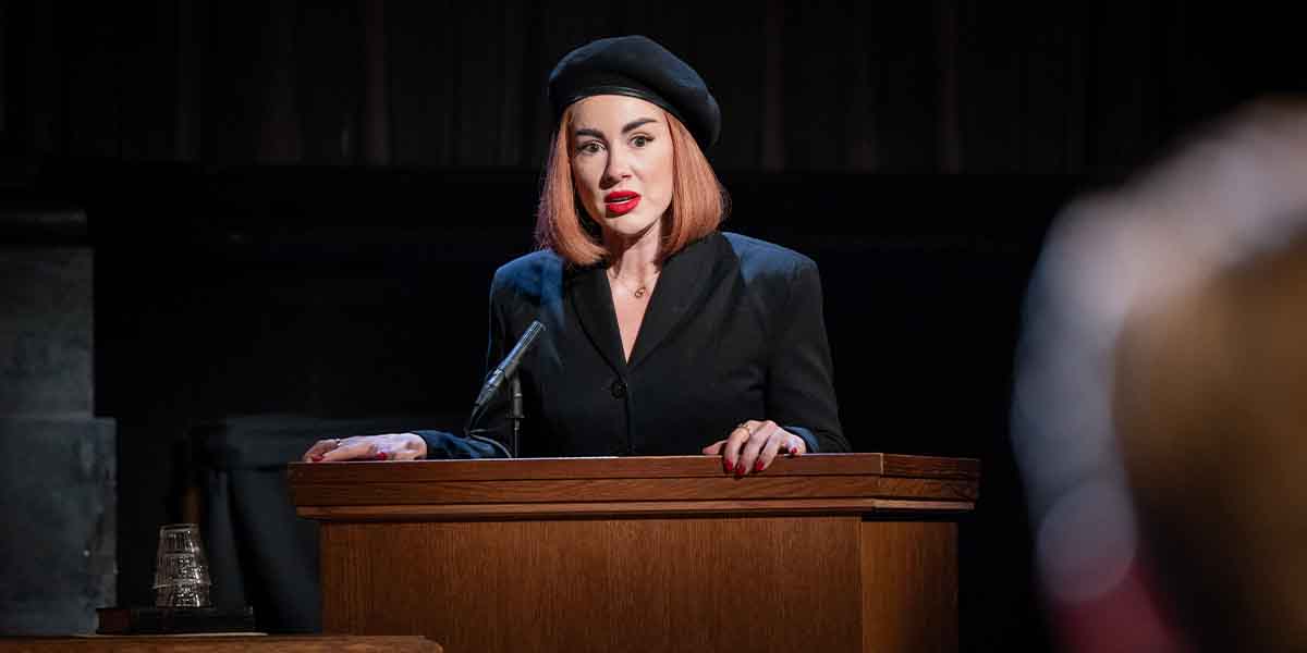 Sophie Craig, as Romaine Vole, stands at a wooden lectern, speaking passionately in a dark setting.