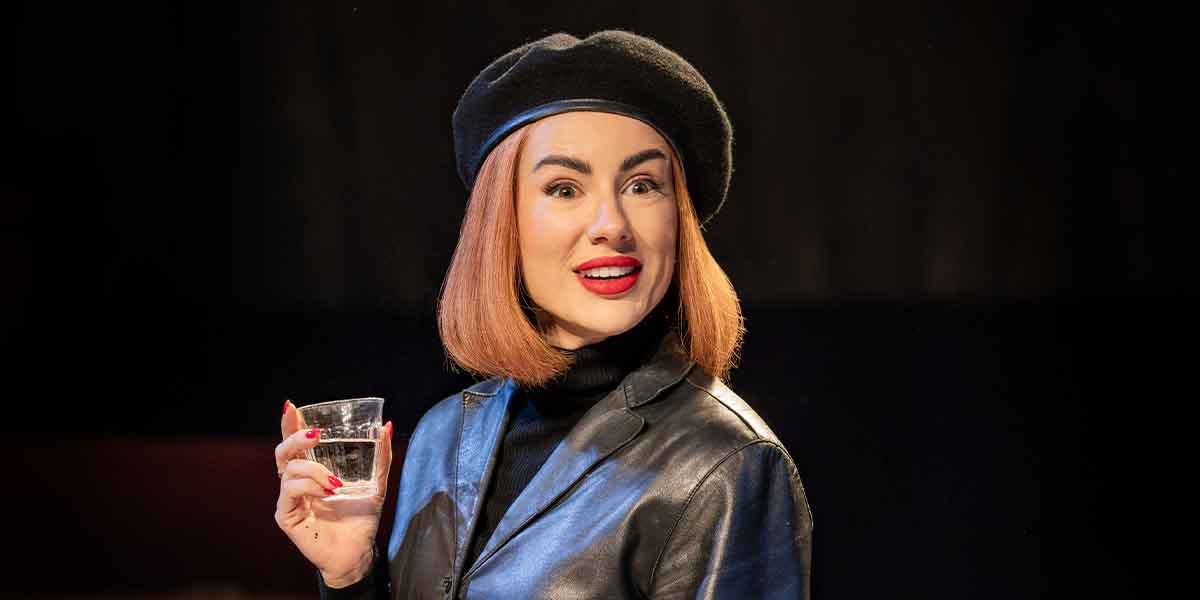 Sophie Craig as Romaine Vole, wearing a black beret and leather jacket, holding a glass of water.