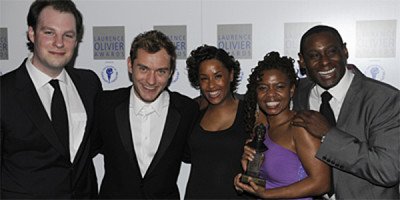 Jude Law poses with two women, one holding an award, against a backdrop of Olivier Awards signage.