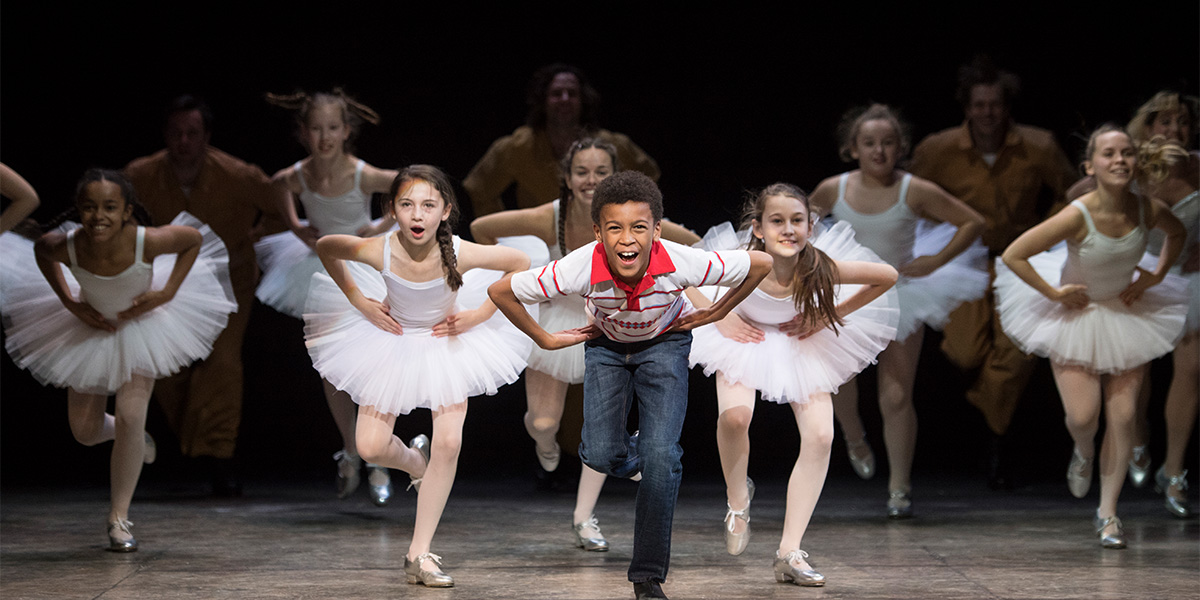 Emile Gooding as Billy Elliot performs with ballet girls, all in colourful costumes, on stage.