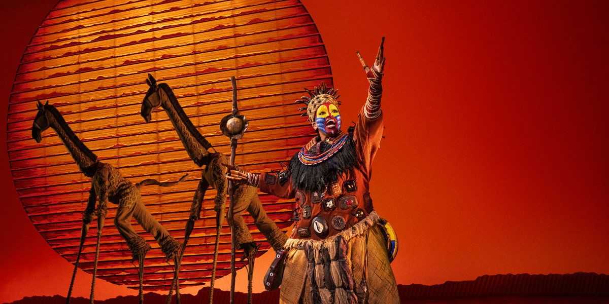A vibrant stage scene from The Lion King, featuring colourful costumes and dramatic lighting in a theatrical setting.