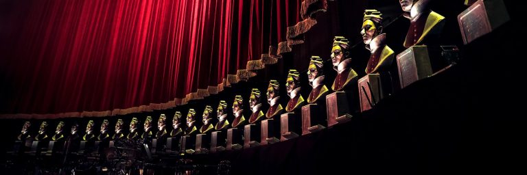 A row of Olivier Award busts against a red velvet curtain, showcasing their distinctive gold and black detailing.