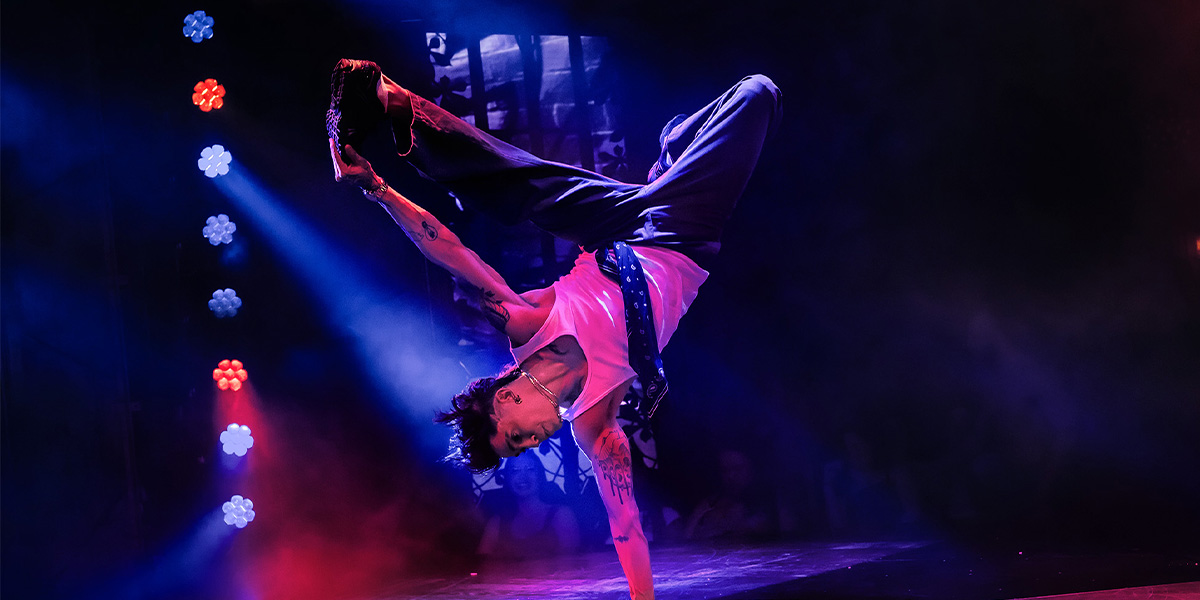 A male performer in a sequined outfit poses confidently on stage, surrounded by dramatic lighting and audience silhouettes.