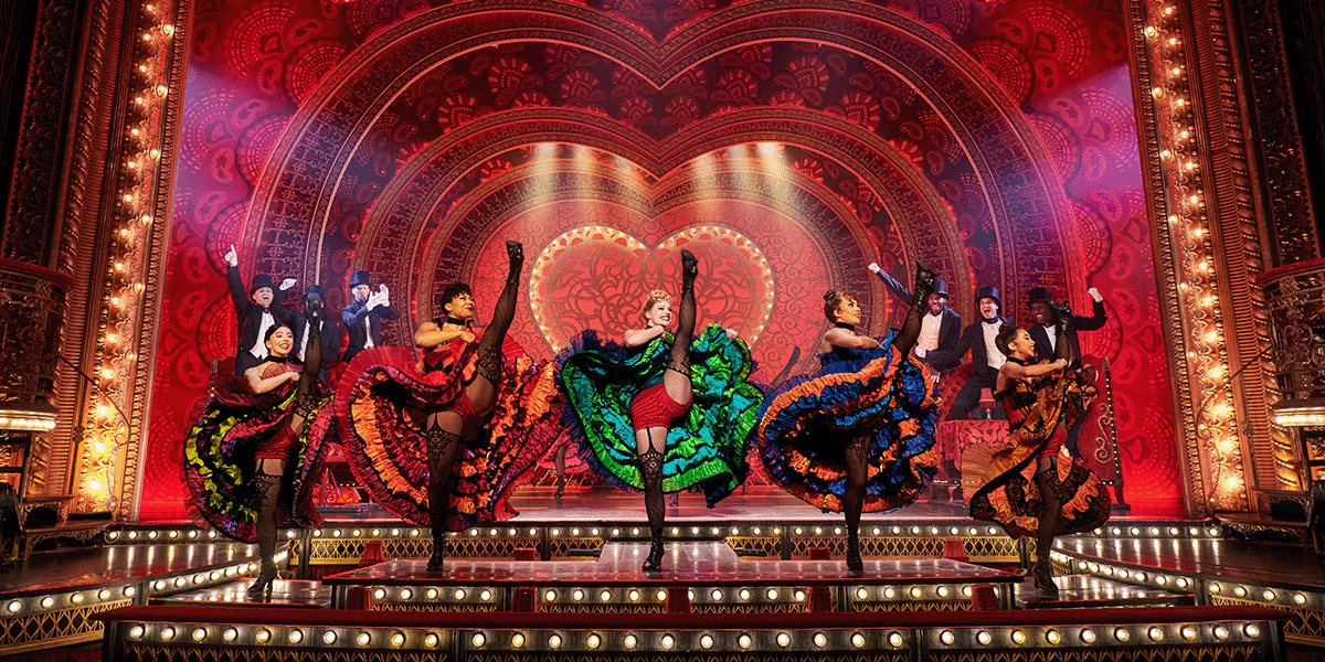 A performer in a red and black costume poses dramatically, surrounded by vibrant stage lights and ornate decor.