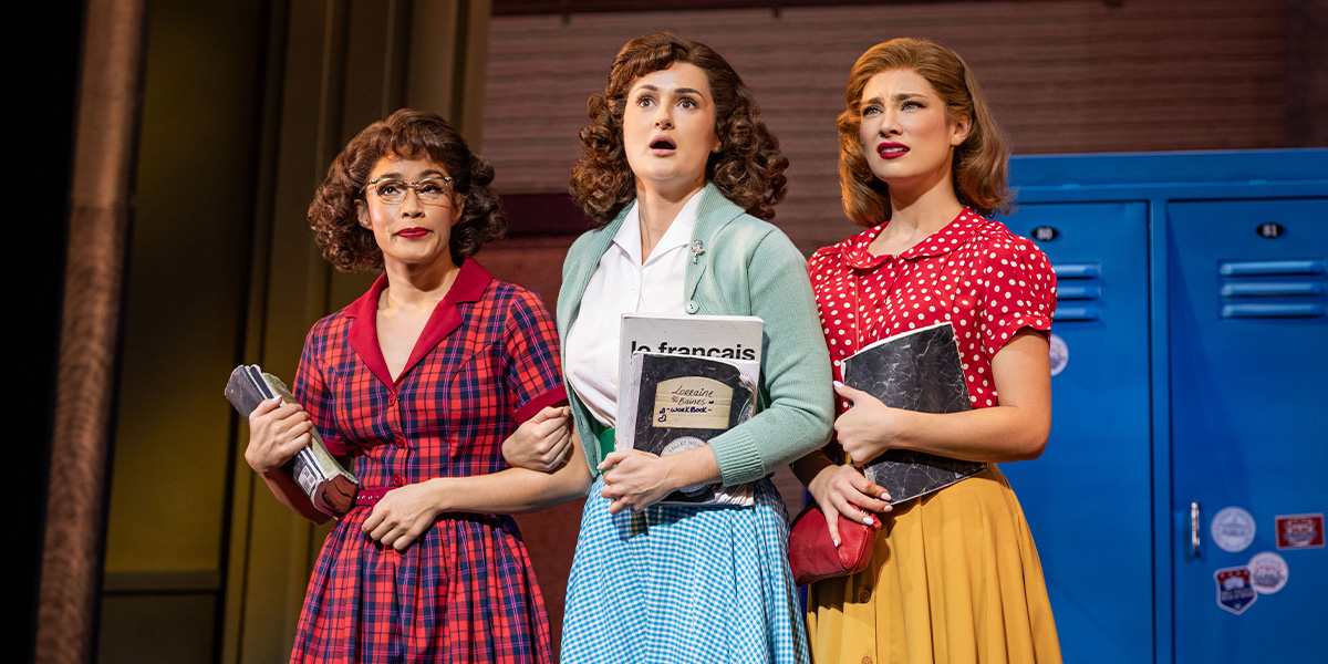 Maddie Grace Jepson as Lorraine Baines stands centre stage, surrounded by cast members in vibrant costumes.