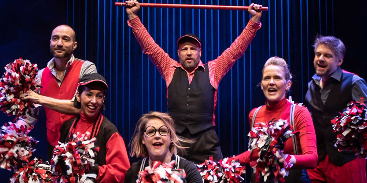 A group of performers in colourful costumes energetically act on stage, with bright lights illuminating the scene.