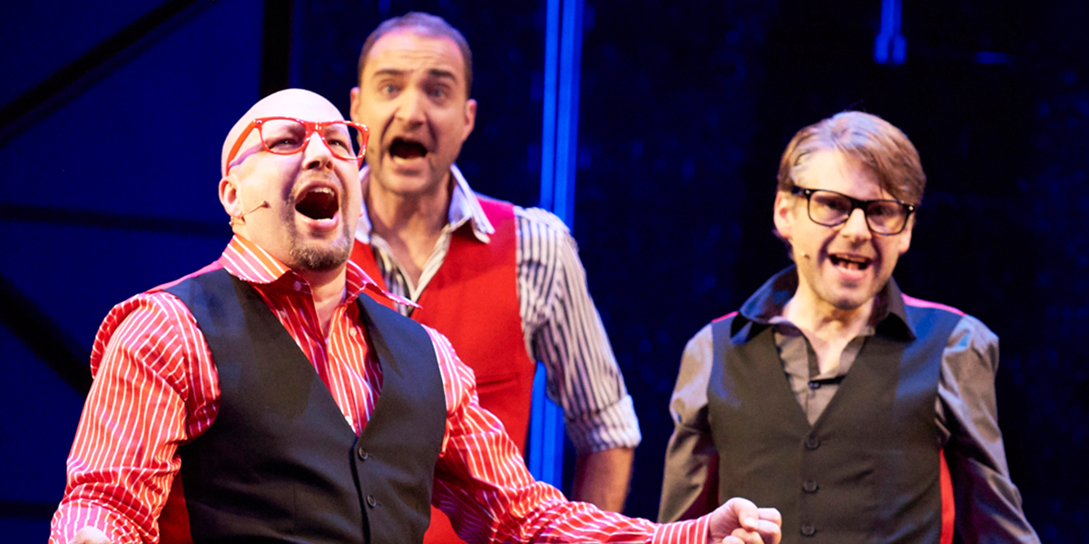 A lively stage scene with performers in colourful costumes, singing and dancing under bright stage lights.