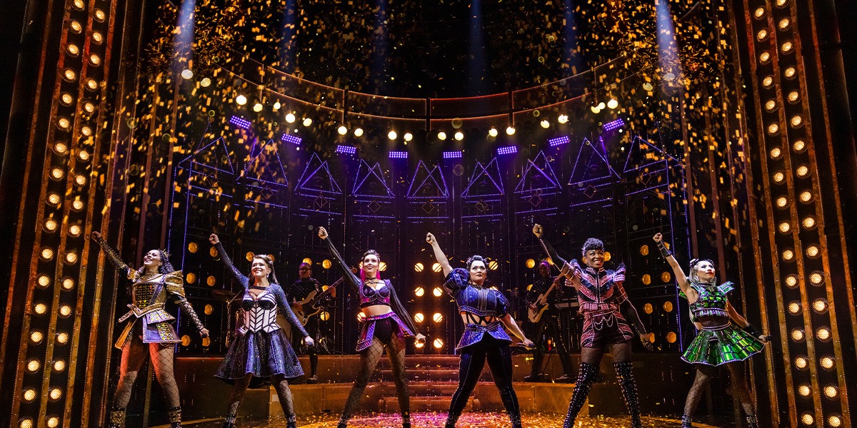 The six performers in vibrant costumes pose confidently, showcasing diverse hairstyles and expressions against a dark stage backdrop.