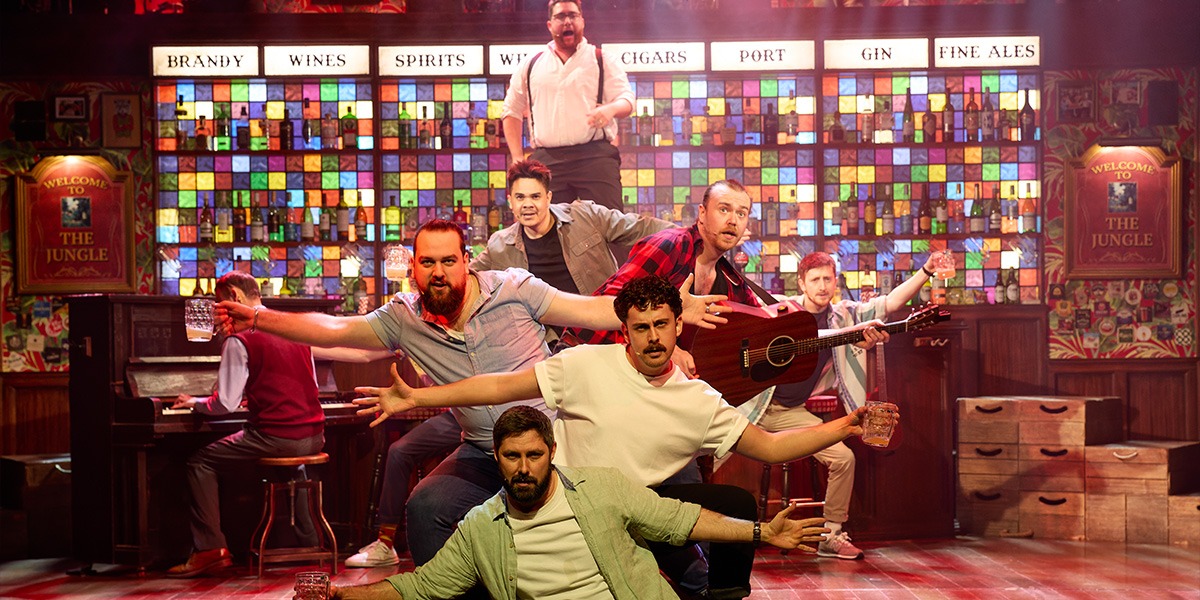 A diverse group of male singers performs energetically on stage, surrounded by musical instruments and warm lighting.