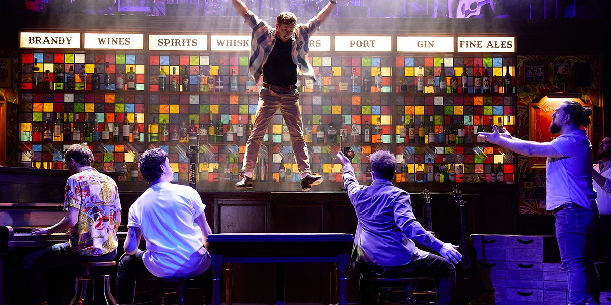 A diverse group of male performers singing energetically on stage, surrounded by bar stools and warm lighting.