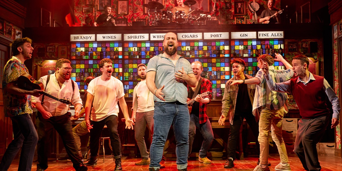 A diverse group of male singers in casual attire perform energetically on stage, surrounded by warm lighting.