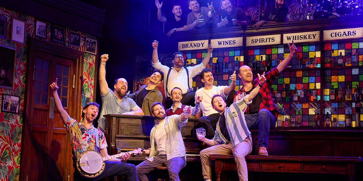 A diverse group of men singing energetically on stage, surrounded by warm lighting and wooden decor.