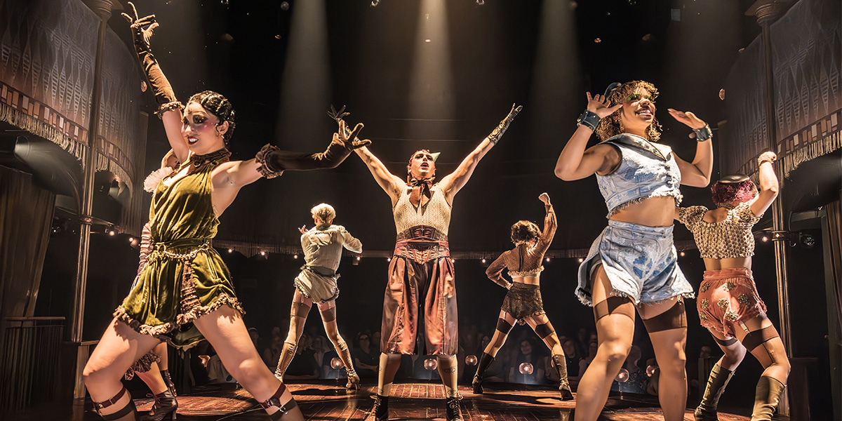 Jamie Muscato as Emcee stands centre stage, surrounded by performers in vibrant costumes, under dramatic lighting.