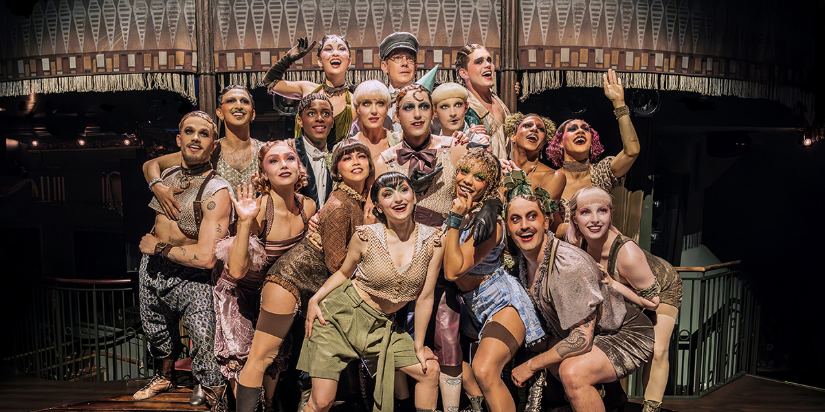 Jamie Muscato as Emcee stands centre stage, surrounded by cast members in vibrant costumes, performing energetically.