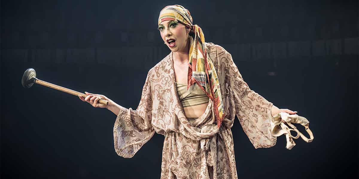 Lindsay Atherton as Fraulein Kost, wearing a patterned robe and headscarf, holds a plunger and gestures.