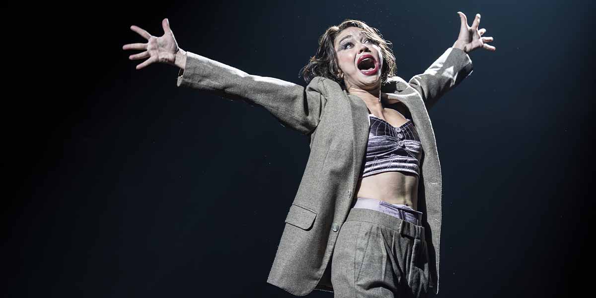 Sarah Hyland as Sally Bowles, arms outstretched, wearing a grey suit and purple top, performing dramatically.