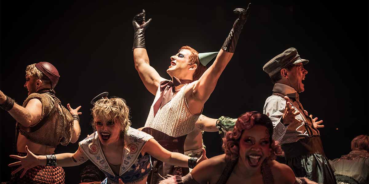 Skylar Astin as Emcee and cast members perform energetically, showcasing vibrant costumes against a dark background.