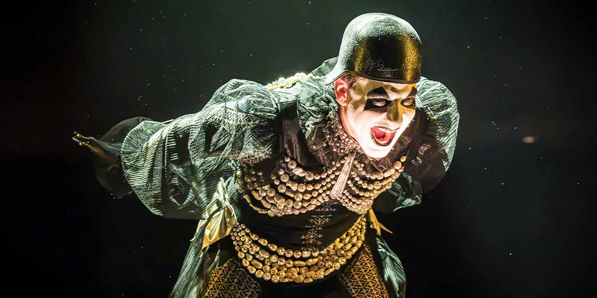 Skylar Astin as Emcee, wearing dramatic makeup and a black costume, passionately performing against a dark background.