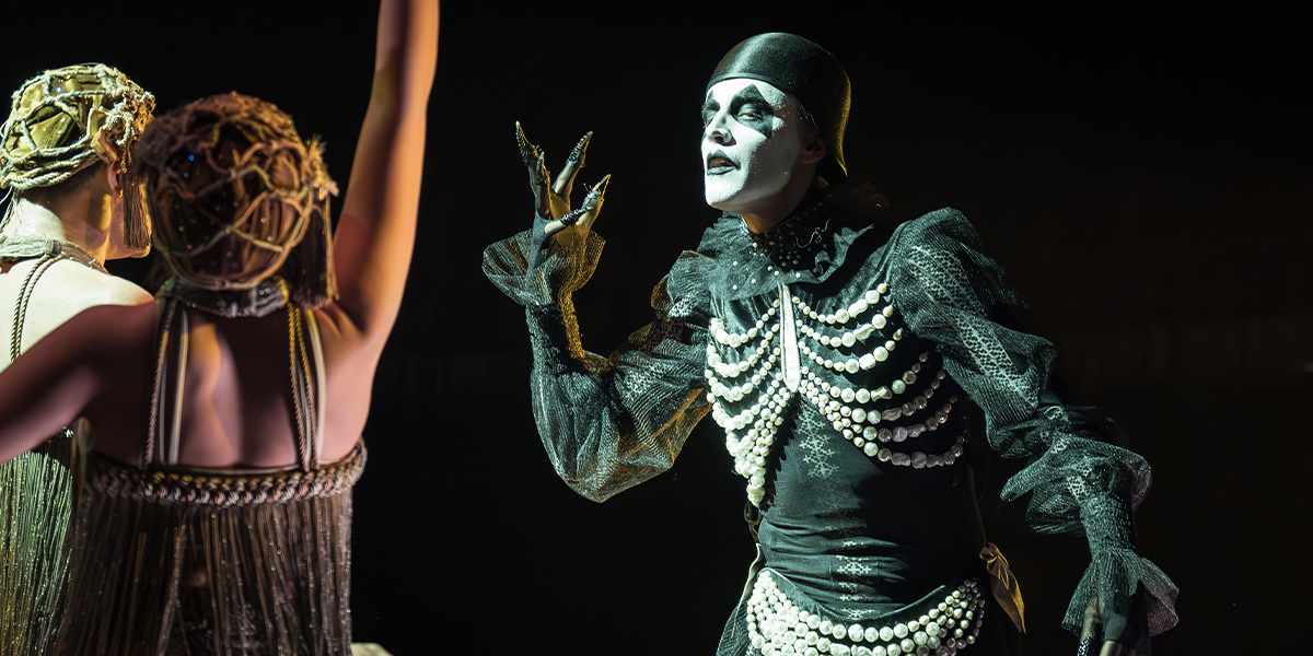 A performer in a sequined costume poses dramatically on stage, with vibrant lighting and a dark backdrop.
