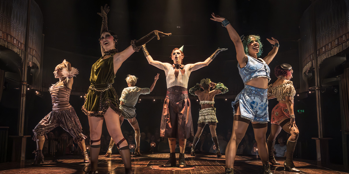 A performer in a sequined costume poses dramatically on stage, with vibrant lighting and a theatrical backdrop.