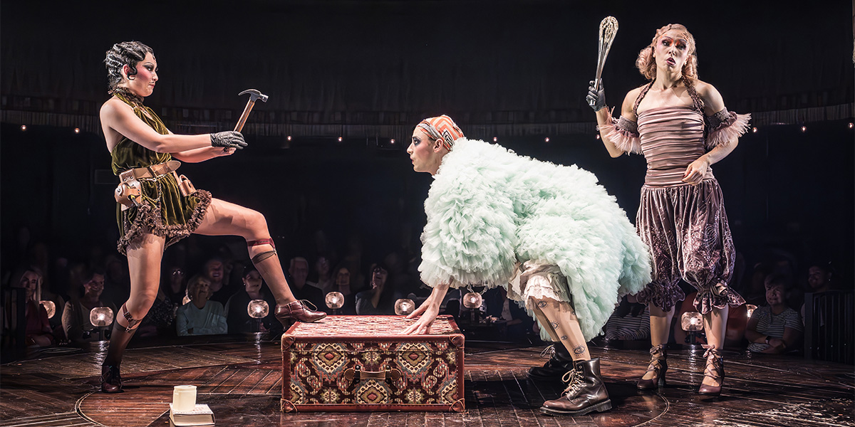 A performer in a sequined costume poses dramatically on stage, with vibrant lighting and a dark background.