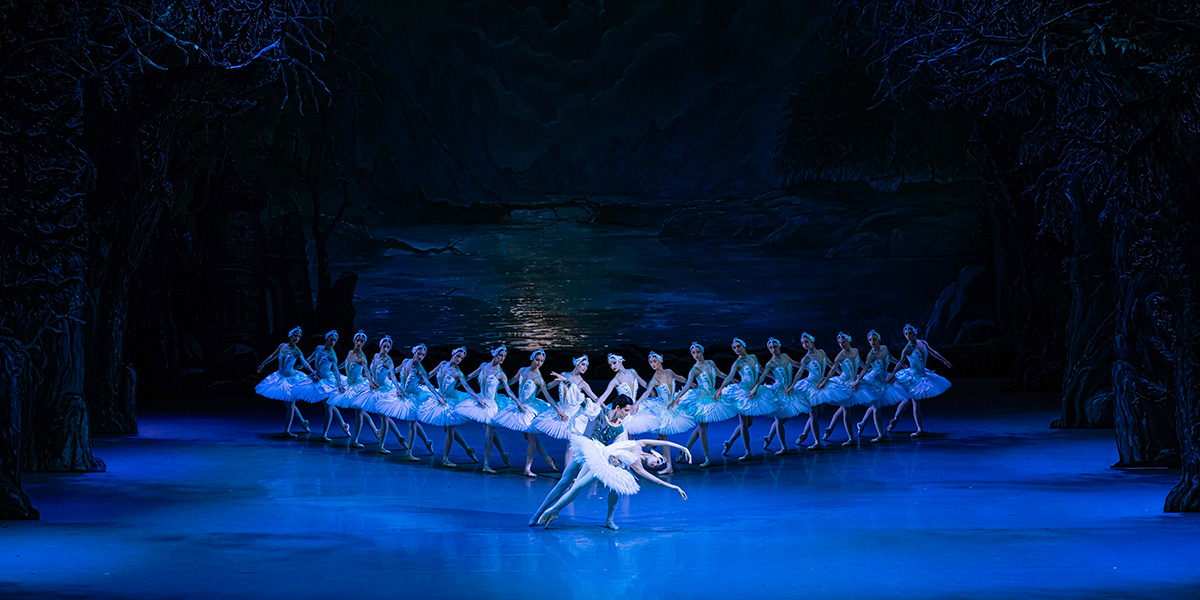 Dancers in elegant white tutus perform a graceful ballet routine on stage, surrounded by soft blue lighting.