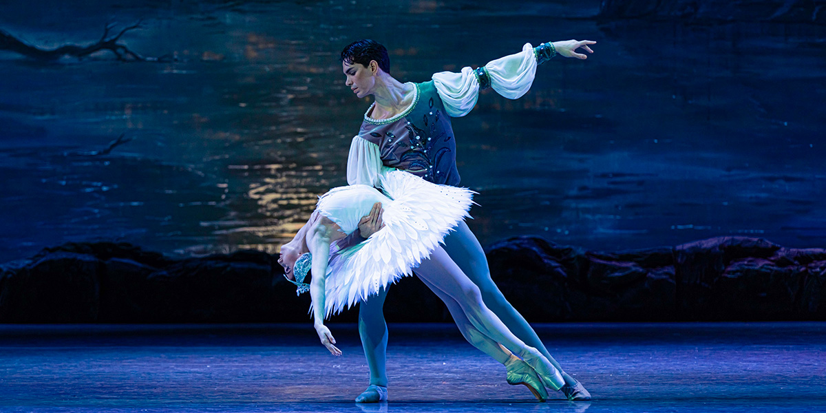 Dancers in elegant white costumes perform a graceful pose, with a dark stage backdrop highlighting their movements.