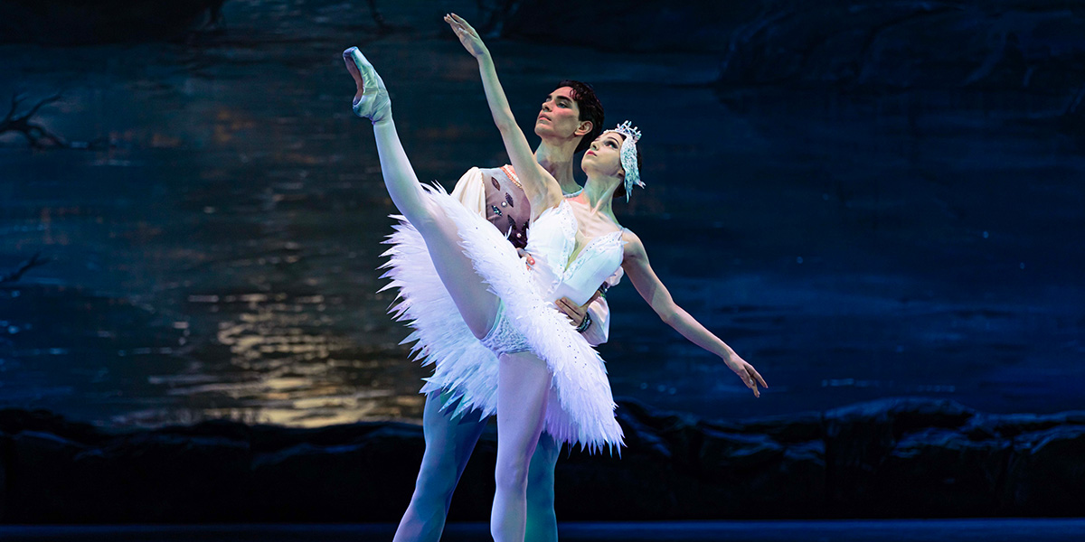 A male dancer lifts a female dancer in a white tutu, set against a dark, serene background.