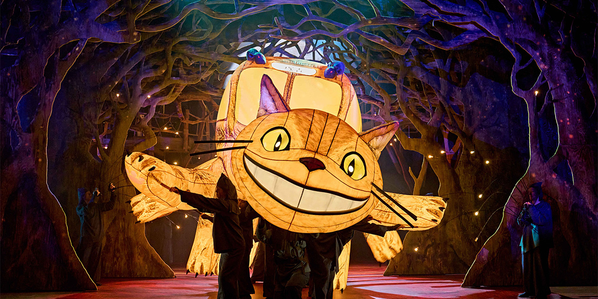 A large, fluffy grey creature with big eyes sits on a grassy hill, surrounded by trees and flowers.