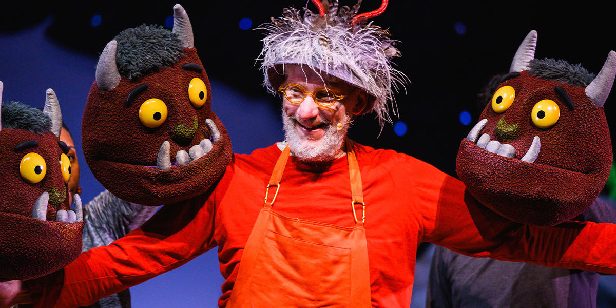 A person in a theater production poses on stage wearing red overalls and spectacles, flanked by three large puppets designed to look like friendly monsters with horns and mischievous smiles.