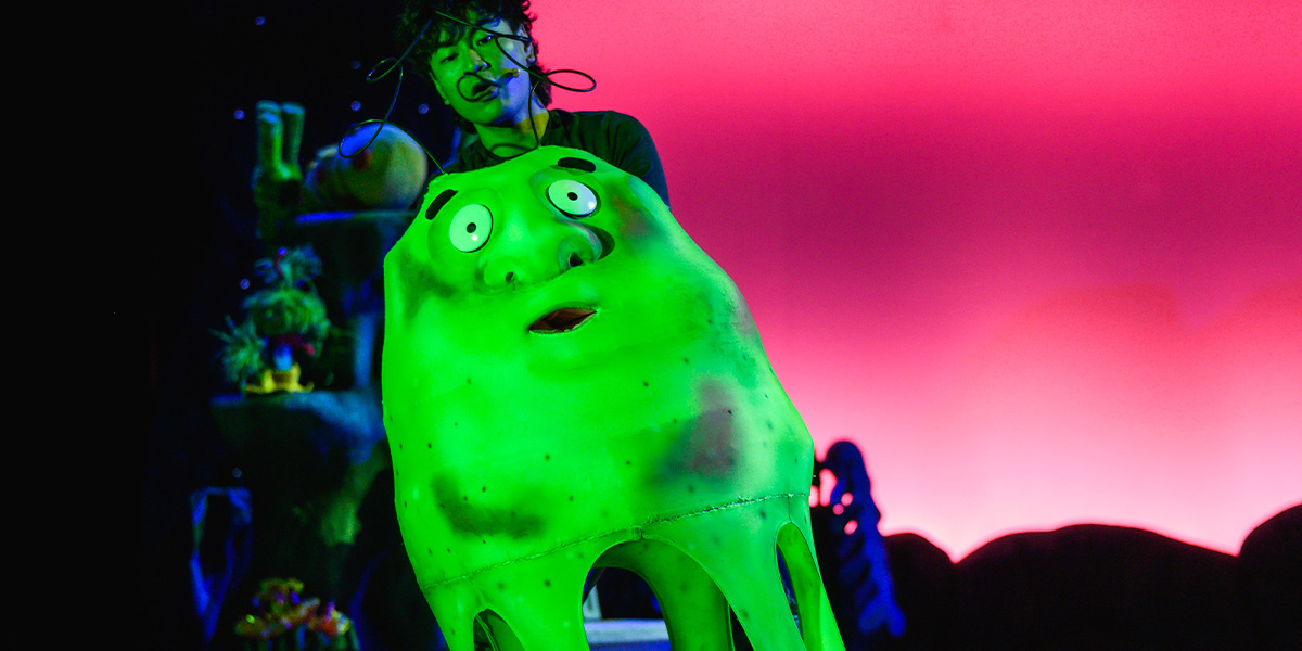 A performer in colourful makeup and costume is engaged dramatically with a large, green, animated character, set against a vibrant red background.
