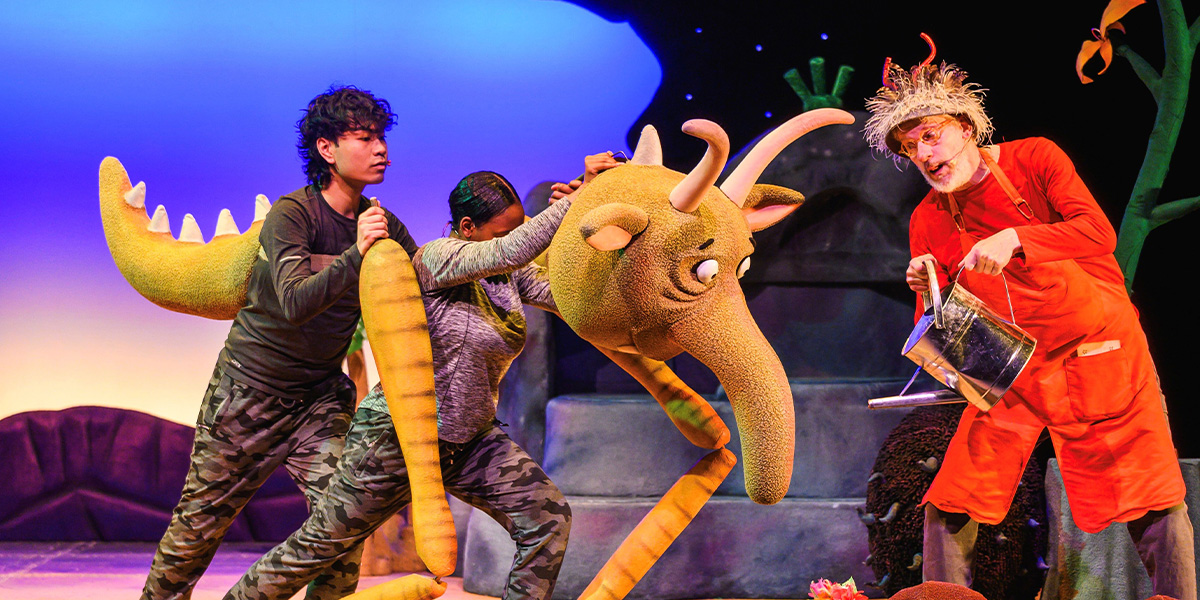 Three actors on stage in a theatrical production, one dressed in a dinosaur costume, another in camouflage, and the third in an orange outfit, interacting dynamically in a colourful set.