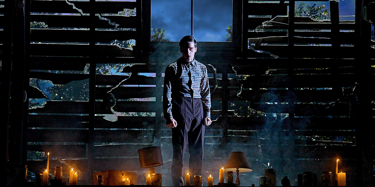 Louis Healy as Henry Creel stands in a dimly lit room, wearing a dark outfit, looking contemplative.