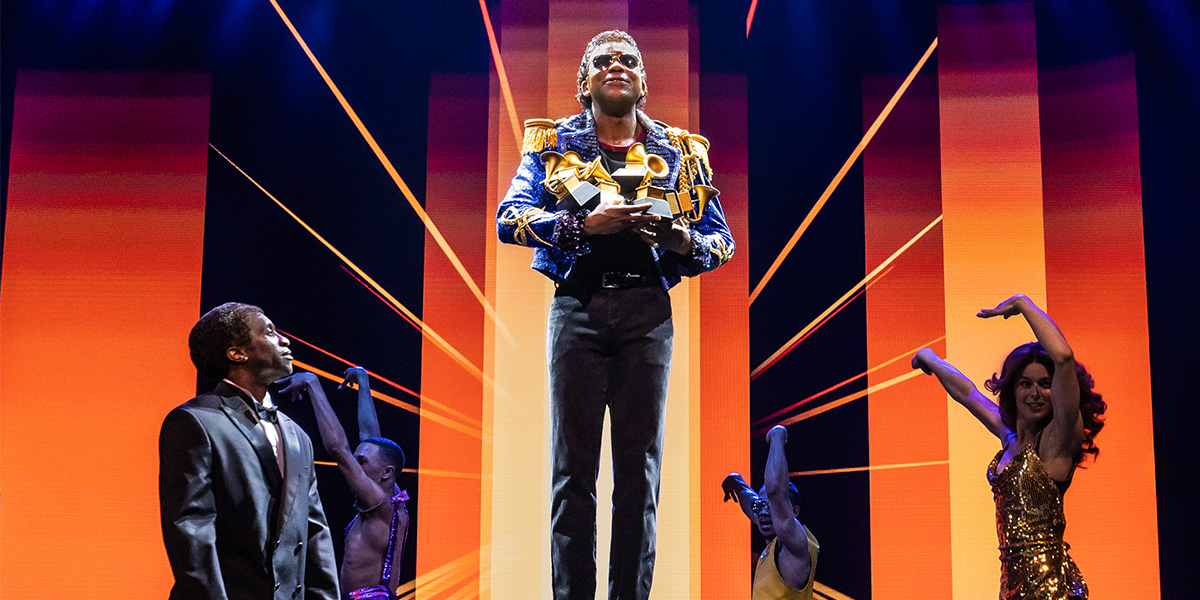 A performer in a sequined costume strikes a dynamic pose on stage, illuminated by vibrant stage lights.