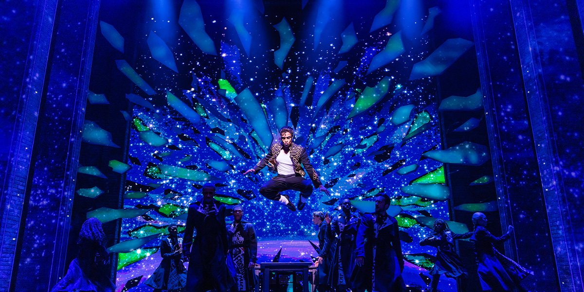 A performer in a sequined jacket gestures energetically on stage, with vibrant stage lights illuminating the background.