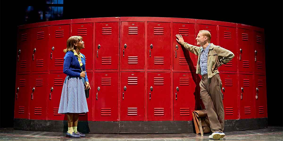 Two characters stand by red lockers, one in a blue sweater and skirt, the other in a checked shirt.