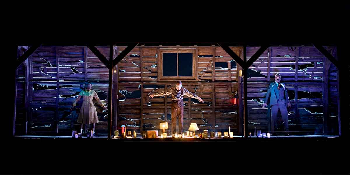 Three characters perform on stage in front of a weathered wooden backdrop, surrounded by candles.