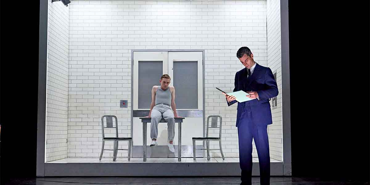 A man in a suit reads a document while a young person sits in a grey outfit behind him.
