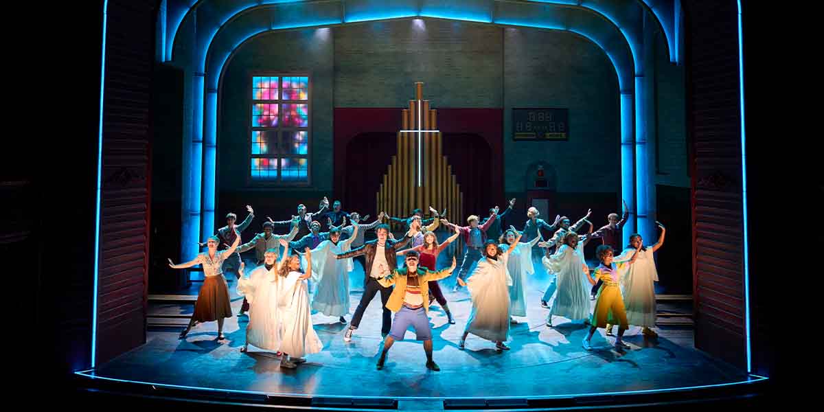A large cast performs on stage, dressed in vibrant costumes, with dramatic lighting and a backdrop.